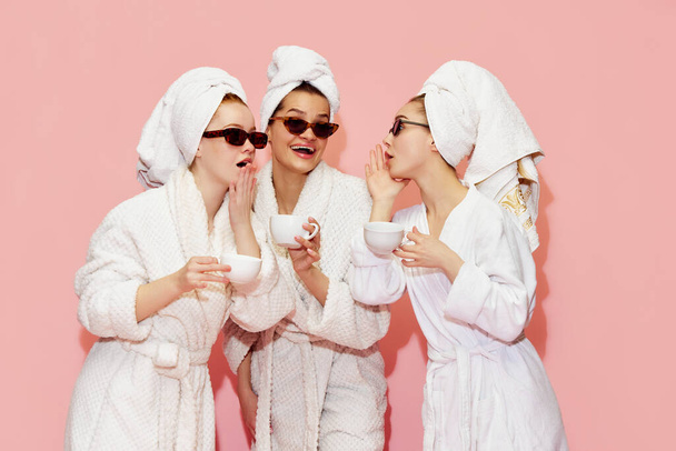 Spreading secrets. Three attractive girls in bathrobes and towels, drinking coffee and talking against pink studio background. Concept of youth, face care, beauty, friendship, party, relaxation - Фото, зображення