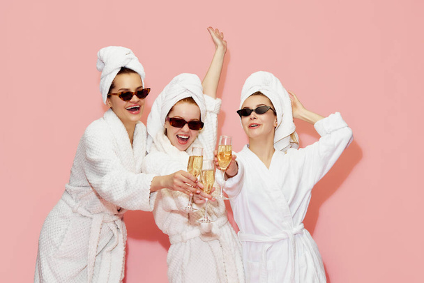 Three beautiful, happy, cheerful girls in bathrobes and towels, drinking champagne, celebrating against pink studio background. Concept of youth, face care, beauty, friendship, hen party, relaxation - Foto, Imagen