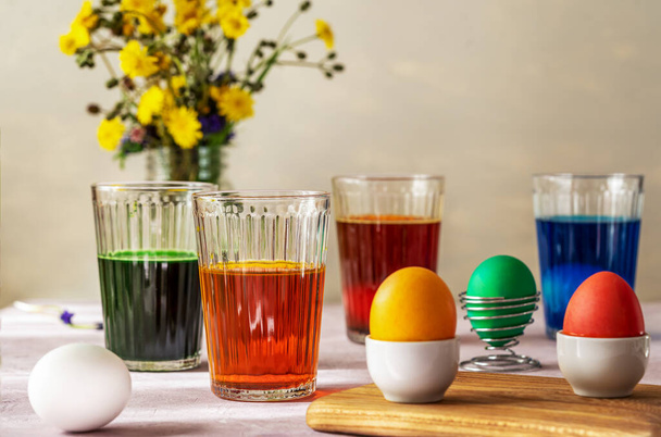 Process of painting Easter eggs. Four glasses with different colours, eggs inside and already painted, wild flowers. Easter celabration concept - Fotó, kép