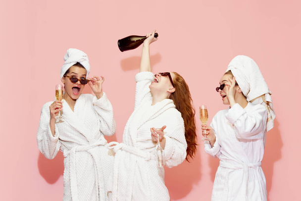 Three happy, beautiful girls in bathrobes drinking champagne, having celebration and fun against pink studio background. Concept of youth, beauty, friendship, party, relaxation, spa - Foto, Bild