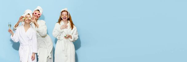 Hen party. Happy, smiling, beautiful girls in bathrobes getting ready, taking care after skin, having fun over blue studio background. Concept of youth, beauty, friendship. Copy space for ad. Banner - Photo, Image