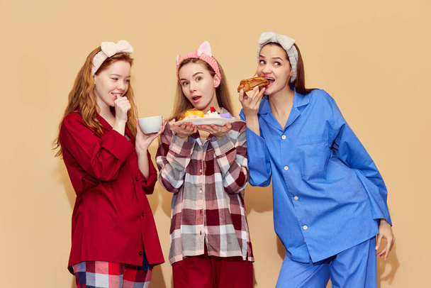 Sleepover. Pretty young girls in colorful cozy pajamas eating sweets, having fun against orange studio background. Concept of youth, emotions, beauty, friendship, party, relaxation - Photo, Image