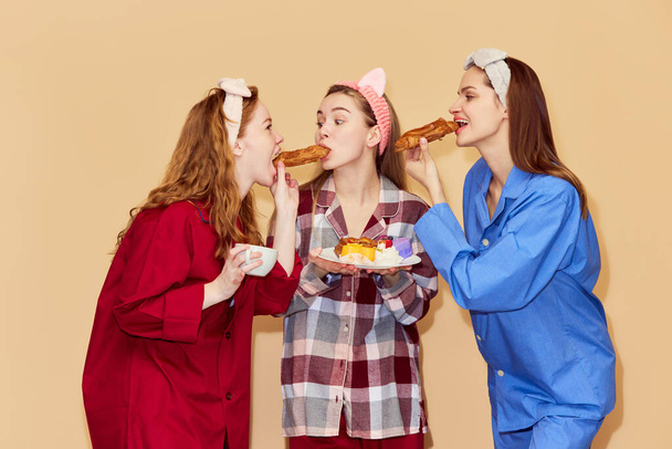 Sweet tooth. Beautiful young girls in comfortable pajamas eating sweets, desserts against orange studio background. Concept of youth, emotions, beauty, friendship, party, relaxation - Photo, image