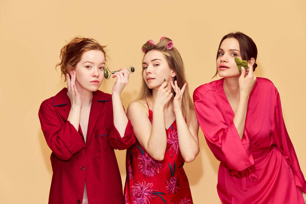 Morning routine. Beautiful young girls in pajamas taking care after skin with cosmetological tools, massaging face against orange studio background. Youth, emotions, beauty, friendship, party concept - Fotó, kép