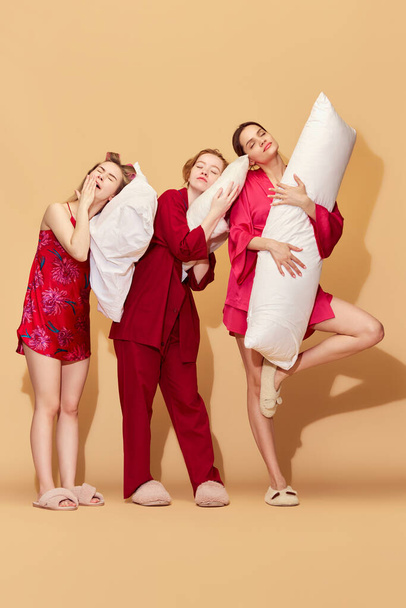 Sleepover with friends. Pretty young girls in pajamas holding pillows and yawning against studio background. Concept of youth, emotions, beauty, friendship, party, relaxation, fun, rest - Foto, Imagem