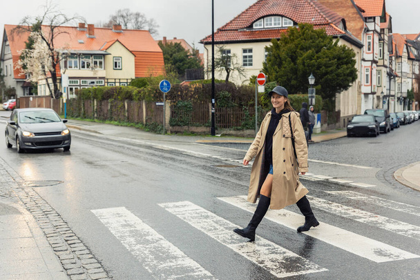 Young adult trendy stylish beautiful caucasian happy smiling woman enjoy walking by road crosswalk in Europe rainy day. Female person portrait wear jeans shorts biege trench coat on urban city street. - Фото, зображення