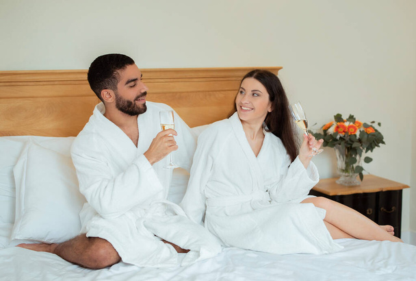 Romantisches multiethnisches Paar trinkt Sekt beim romantischen Date im Hotel. Ehepartner genießen Champagner im Bett in der Luxus-Suite im Flitterwochen-Urlaub und tragen weiße Bademäntel - Foto, Bild