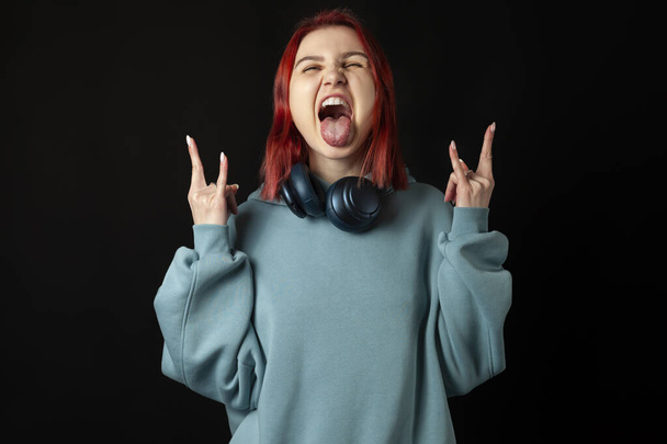 Cool young redhead girl in a blue oversized hoodie shouting with crazy expression doing rock symbol with hands up. Music star. Heavy concept. Sign of the horns, tongue is out. isolated on black. - Photo, Image