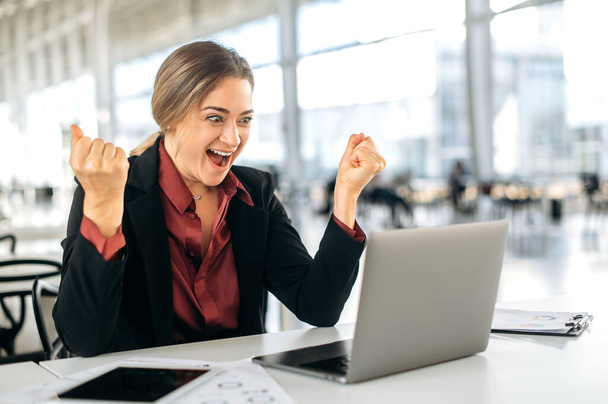 Joyful excited caucasian woman, manager, office employee, sits at a work desk in office, looks in amazement at the laptop screen, rejoices at success, good result, news, gesticulates with her hands - Foto, imagen