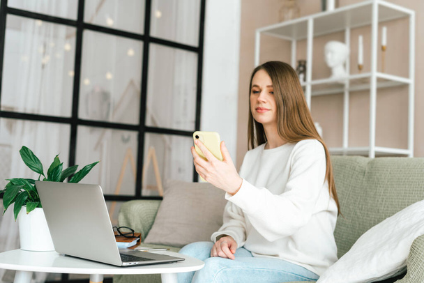 Freelance young woman with mobile cellphone typing at laptop and working from home office with. Happy girl on workplace at desk. Distance learning online education. Taking selfie or video chat - Photo, Image