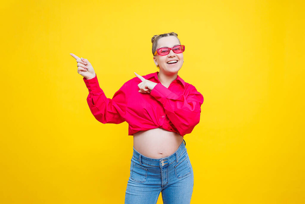 Gelukkige zwangerschap. Vrolijke mooie zwangere vrouw in een roze shirt en roze bril op een gele achtergrond. Jonge heldere zwangere vrouw. - Foto, afbeelding