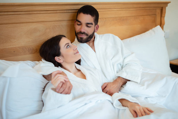 Διαφορετικό ερωτευμένο ζευγάρι ξαπλωμένο στο κρεβάτι στο εσωτερικό της κρεβατοκάμαρας, φορώντας λευκά μπουρνούζια. Σύζυγοι κοιτάζονται χαλαρώνοντας στο ξενοδοχείο Σουίτα για διακοπές. Ρομαντικό μήνα του μέλιτος - Φωτογραφία, εικόνα
