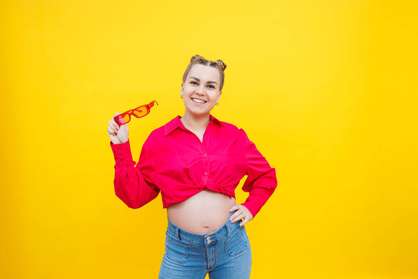 Gelukkige zwangere vrouw met een glimlach op haar gezicht. Vrolijke mooie zwangere vrouw in een roze shirt en roze bril op een gele achtergrond. Jonge heldere zwangere vrouw. - Foto, afbeelding