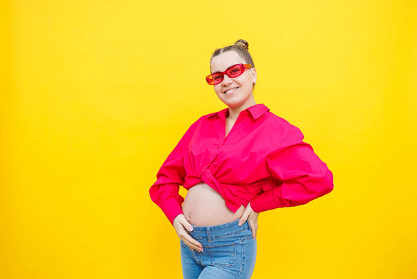 Boldog terhes nő mosollyal az arcán. Vidám, csinos, terhes nő rózsaszín ingben és rózsaszín szemüvegben, sárga háttérrel. Fiatal okos terhes nő. - Fotó, kép