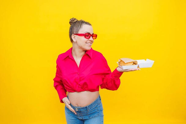 Una mujer embarazada con una camisa rosa y jeans sostiene una sabrosa hamburguesa. Alimento nocivo durante el embarazo. Concepto de embarazo fácil y feliz. - Foto, Imagen