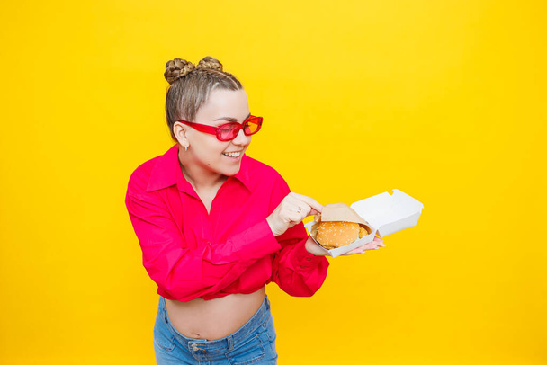 Pembe gömlekli ve kotlu hamile bir kadın lezzetli bir burger tutuyor. Hamilelik boyunca zararlı yiyecekler. Kolay ve mutlu hamilelik kavramı. - Fotoğraf, Görsel