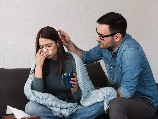 Young couple sitting on sofa, woman is covered with blanket, coughing and blowing nose. Cold and flu season - Photo, Image