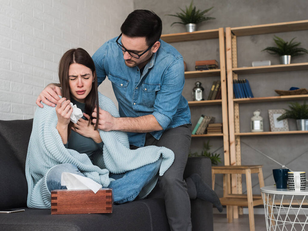 Young couple sitting on sofa, woman is covered with blanket, coughing and blowing nose. Cold and flu season - Photo, Image
