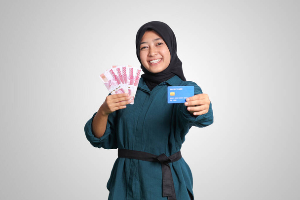 Portret wesołej muzułmanki azjatyckiej z hidżabem, pokazujący sto tysięcy rupii trzymającej kartę kredytową. Koncepcja finansów i oszczędności. Izolowany obraz na białym tle - Zdjęcie, obraz