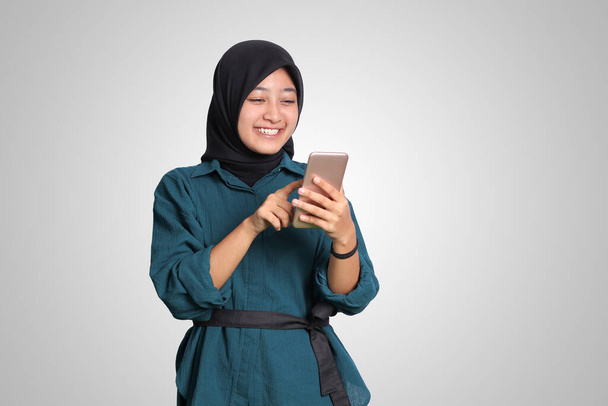 Heyecanlı Asyalı Müslüman kadının portresi tesettür tutan ve cep telefonuna dokunan. Reklam konsepti. Beyaz arkaplanda izole edilmiş resim - Fotoğraf, Görsel