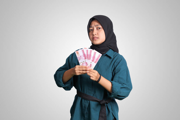 Portret podekscytowanej azjatyckiej hidżabki w luźnym stroju pokazujący sto tysięcy rupii. Koncepcja finansów i oszczędności. Izolowany obraz na białym tle - Zdjęcie, obraz