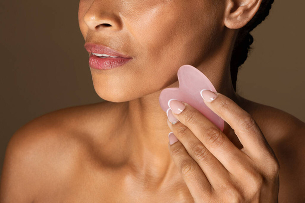 Cosmetic natural trend. African american woman using beauty Gua Sha quartz stone for massage, standing on brown background, closeup. Traditional medicine and cosmetology concept. - Photo, Image