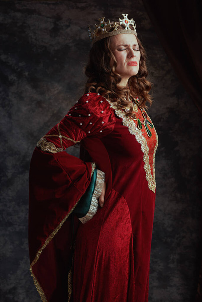 tired medieval queen in red dress with crown having backache on dark gray background. - Photo, Image