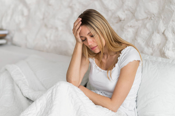 Отчаянная кавказская блондинка, сидящая на кровати, сжимает руку к голове, страдает мигренью и болезнью в интерьере белой спальни. Симптомы простуды и гриппа, головная боль, проблемы со здоровьем, депрессия - Фото, изображение