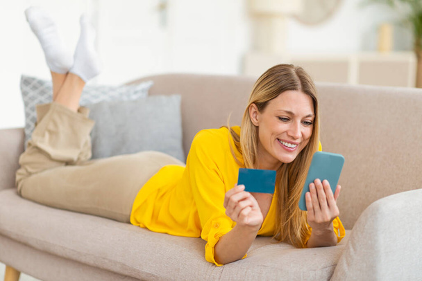 Banking app, pay money. Smiling millennial caucasian blonde woman shopaholic in yellow clothes with credit card and smartphone shopping online in living room interior. Sale, check financial account - Fotoğraf, Görsel