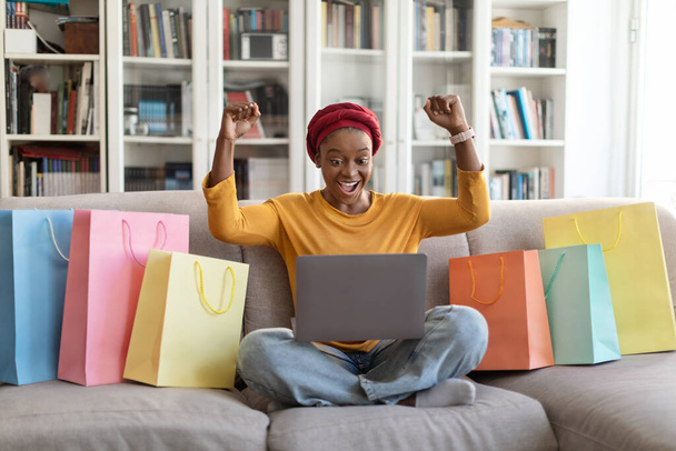 Black friday, sales. Happy excited pretty young black woman in red turban shopaholic sitting on couch with lot of colorful shopping bags, using laptop at home, raising hands up, copy space - Photo, Image