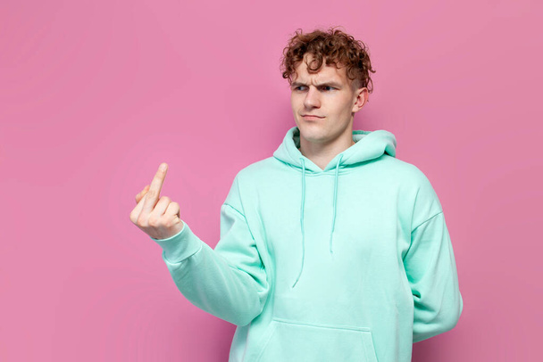 young rude guy in mint hoodie showing middle finger on pink isolated background, furious man showing fuck with his hands - Photo, image