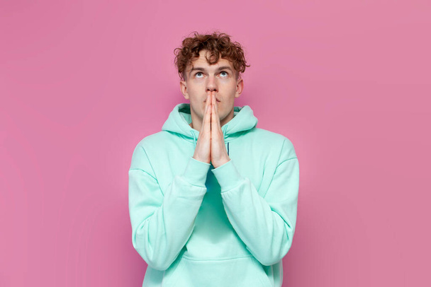 young curly guy in mint hoodie pray and hope for good luck on pink isolated background, man folds his hands in front of him and shows gesture of faith - Foto, Imagem