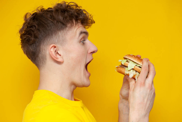 young curly guy holding a burger with his mouth open on a yellow background, a man eats fast food, close-up - Zdjęcie, obraz