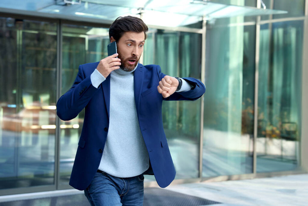 Emotional businessman talking on smartphone and running near building. Being late concept - Photo, image
