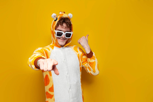 jeune homme joyeux en pyjama girafe drôle d'enfants et des lunettes vous invite à faire la fête sur fond jaune, l'homme en vêtements de cosplay animal montre sa main à vous, le concept de fête pyjama - Photo, image