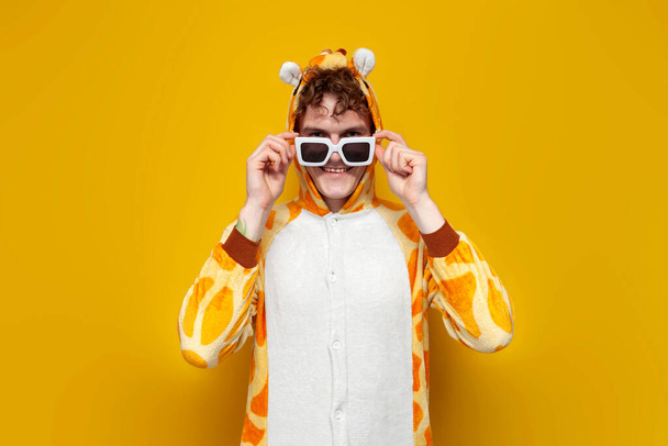 młody radosny facet w zabawnej piżamie i okularach dziecięcej żyrafy na żółtym tle, mężczyzna w ubraniu dla zwierząt cosplay, koncepcja piżamy party - Zdjęcie, obraz