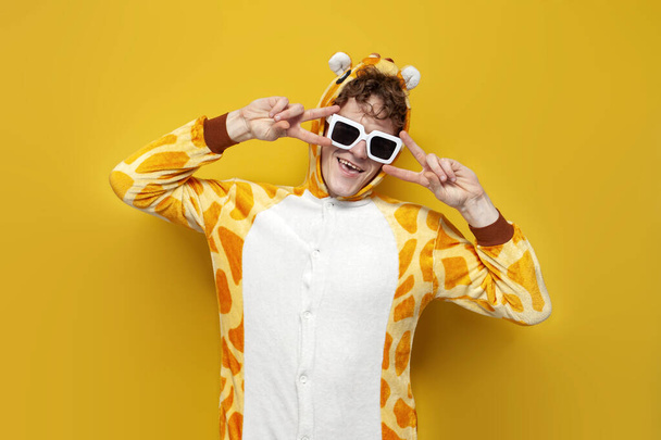 joven alegre chico en divertido bebé jirafa pijamas y gafas está bailando sobre fondo amarillo, hombre en animal cosplay ropa muestra gesto de paz, concepto de fiesta de pijama - Foto, Imagen