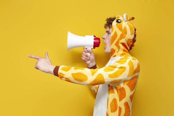 young joyful guy in funny children's giraffe pajamas speaks into megaphone and points his hand to the side on yellow background, man in animal cosplay clothes announces and shouts - Photo, Image