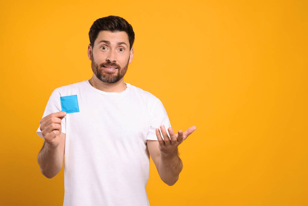 Confused man holding condom on yellow background, space for text. Safe sex - Photo, image