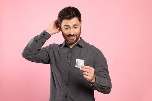 Confused man holding condom on pink background. Safe sex - Photo, Image