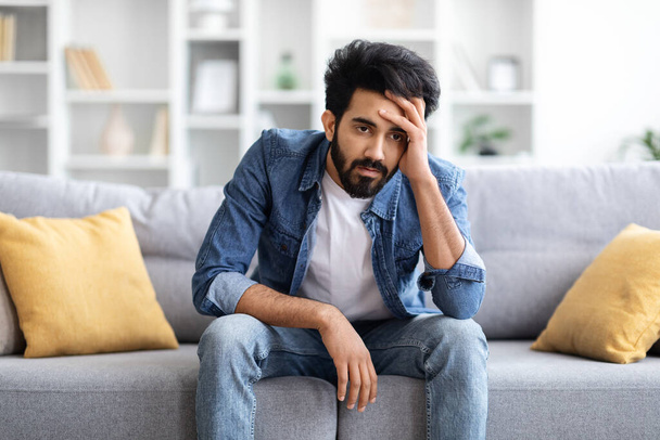 Portrait Of Depressed Young Indian Man Sitting On Couch At Home, Pensive Upset Eastern Guy Touching Head And Looking Away, Suffering Depression, Life Problems Or Mental Breakdown, Copy Space - Photo, Image