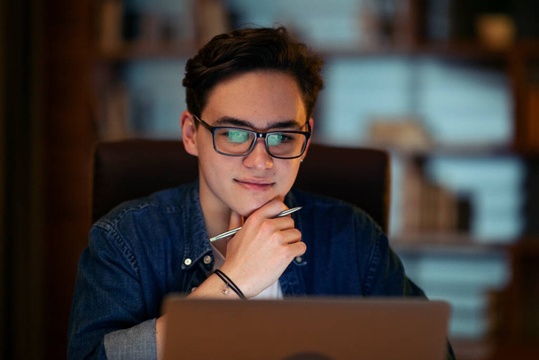 Closeup of happy young man wearing eyeglasses working overtime alone at his desk in office late at night, looking at laptop screen and smiling, holding stylus pen in his hand, copy space - Foto, immagini