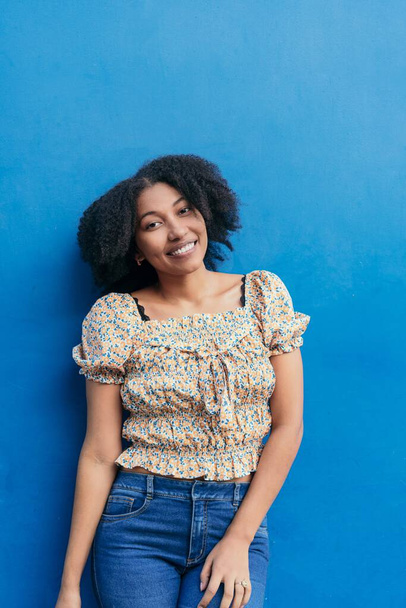 Αφρο-Αμερικανίδα με όμορφα αφρο μαλλιά. Κοιτάζει την κάμερα με μια χαμογελαστή έκφραση σε ένα μπλε φόντο.. - Φωτογραφία, εικόνα