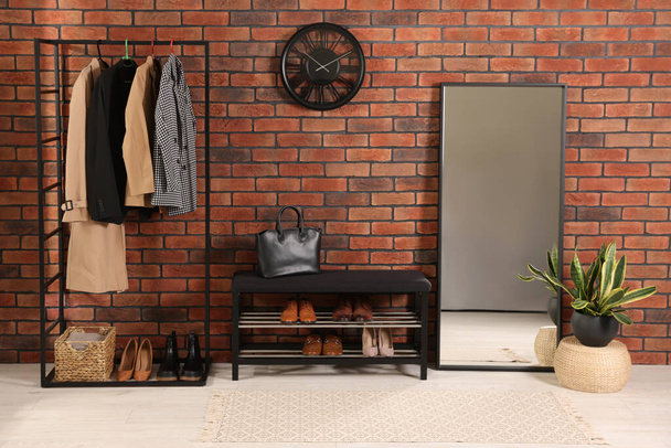 Stylish hallway with coat rack, mirror and shoe storage bench near brick wall. Interior design - Zdjęcie, obraz