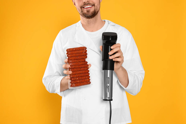 Smiling chef holding sous vide cooker and sausages in vacuum pack on orange background, closeup - Фото, зображення