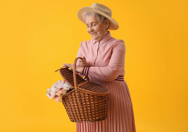 Ηλικιωμένη γυναίκα με καλάθι με λουλούδια σε κίτρινο φόντο - Φωτογραφία, εικόνα