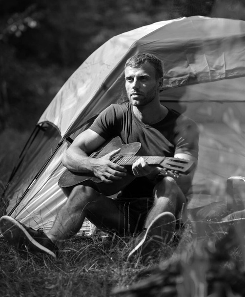 Young man sitting near touristic tent and playing guitar. Romantic time in a mountain camp. Tourism and hike, travel lifestyle. Handsome attractive model man playing guitar in forest with bonfire - Fotografie, Obrázek