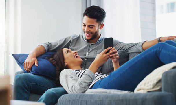 Passons en revue nos photos. un jeune couple affectueux se prélassant sur le canapé tout en utilisant un téléphone portable dans leur salon - Photo, image