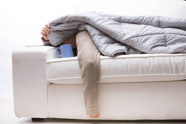 woman, lying on front, white couch, warm quilt, sweater, overwhelmed, flu, peeking, lifting fabric, weak arm, exhausted, hot tea, mug, sickness, illness, influenza, covered, rest, recovery, fatigue, t - Фото, изображение
