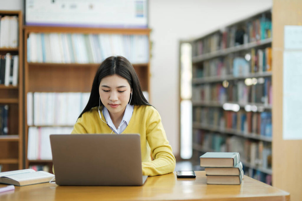 Young female college, highschool student wearing headphones and casual clothings sitting at desk reading book, studying and doing research using laptop for school project at a library. E-Learning Educational Library concept. - 写真・画像